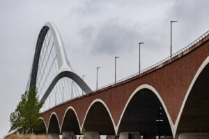A striking modern arch bridge with brick arches and metal curves under a cloudy sky.