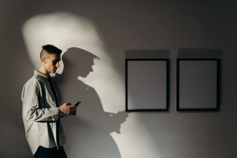 A man holding a smartphone stands indoors against a minimalistic, shadowed wall featuring blank frames.