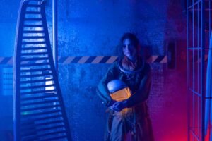 A woman in a spacesuit holding a helmet inside a sci-fi setting with neon lights.