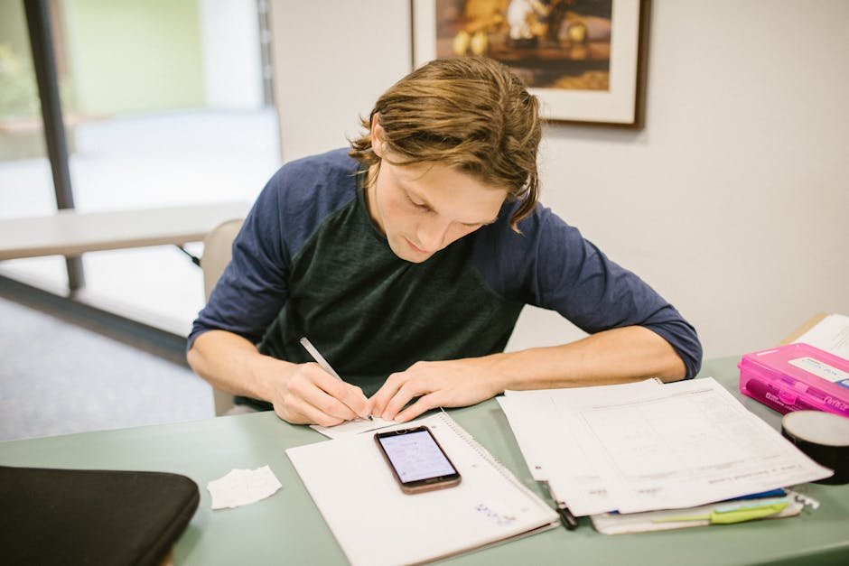 Male college student writing notes at a desk with a smartphone during study session.