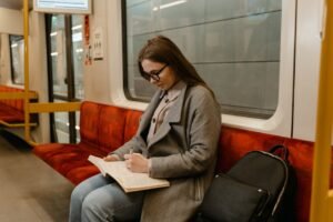 A woman in glasses and a gray coat writes in a notebook while commuting on a train.